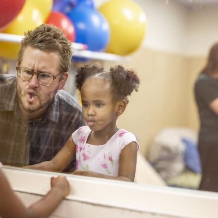 A white male therapist with glasses and young girl with brown skin and pigtails make "fish faces" in a mirror.
