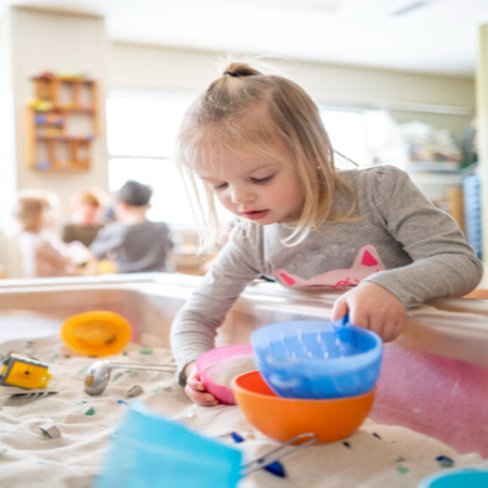 Blonde preschooler with a ponytail calmly playing at a sensory table filled with sand and toys.