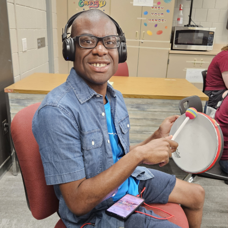 A smiling dark-skinned boy with glasses, wearing headphones, banging on a drum.
