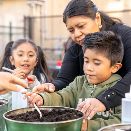 Tan-skinned mother and two children planting herbs at an outdoor event. 