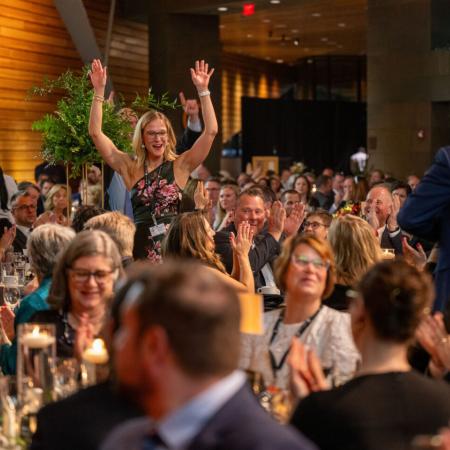 Woman raises her hands in the air in victory as she walks up to receive her raffle prize. She is surrounded by people at dinner tables applauding.