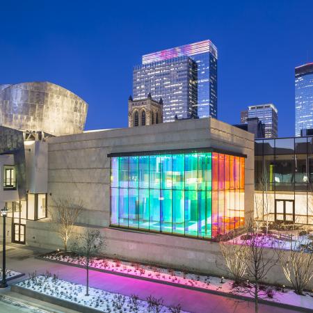 A nighttime photo of part of the downtown Minneapolis skyline featuring a building with internally lit windows casting a rainbow glow onto the sidewalk and street. 
