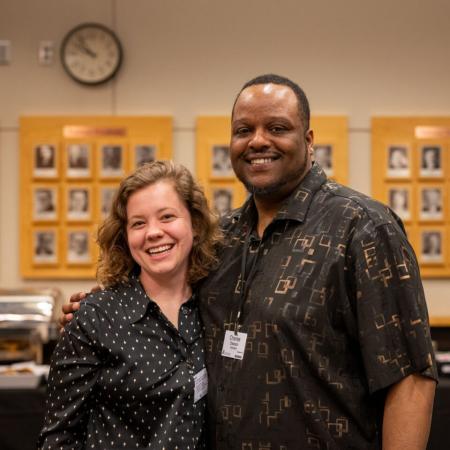 A white woman with curly hair wearing a dark shirt, smiling next to a dark-skinned man wearing a dark shirt. 