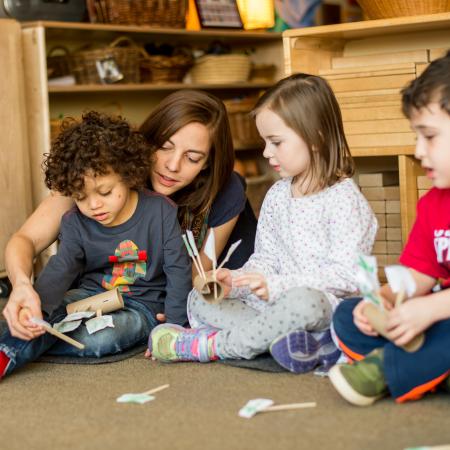 A teacher sits on the floor and interacts with three children, in a classroom.