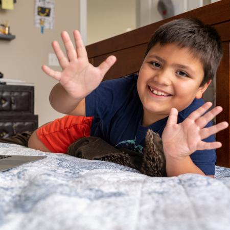 Little boy with short brown hair sits on a bed next to a laptop showing his therapist during a telehealth session.