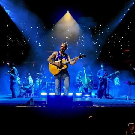 Zach Bryan holding a guitar on stage, singing, with a band behind him in bluish light.