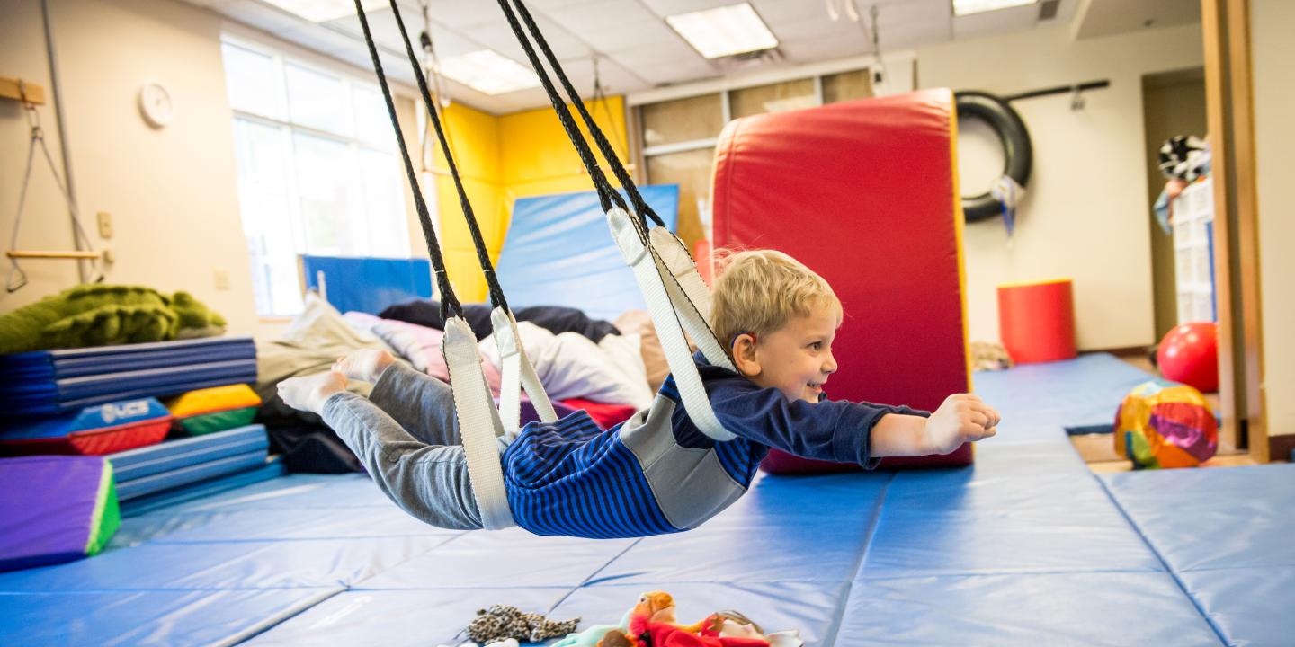 Little boy making a superman pose as he swings on his stomach, on a therapy swing.