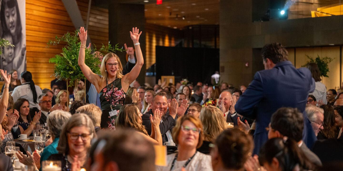 Woman raises her hands in the air in victory as she walks up to receive her raffle prize. She is surrounded by people at dinner tables applauding.