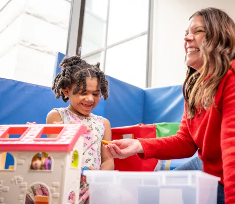 A young girl with braids plays with a toy house with a smiling female therapist wearing a bright red sweater.