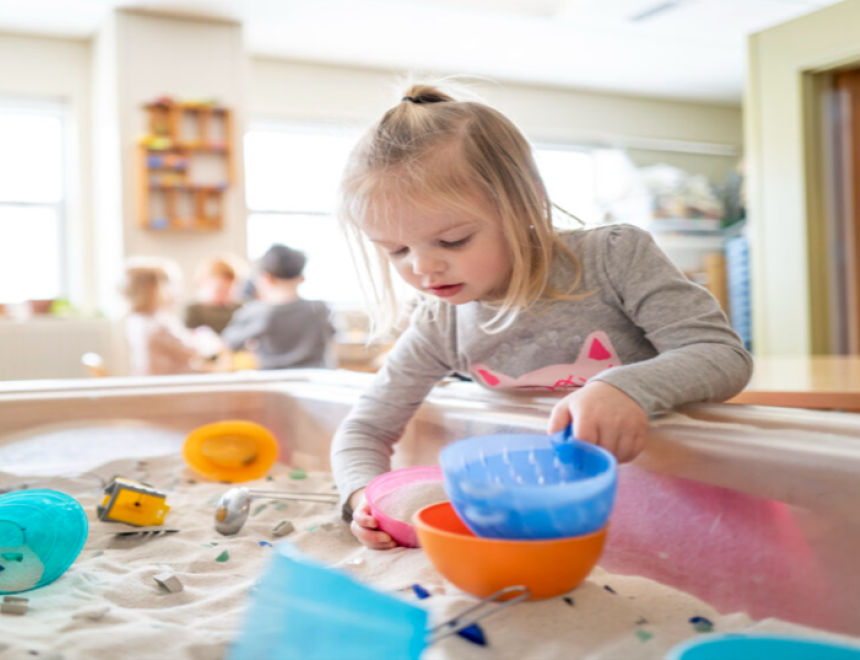 Blonde preschooler with a ponytail calmly playing at a sensory table filled with sand and toys. 
