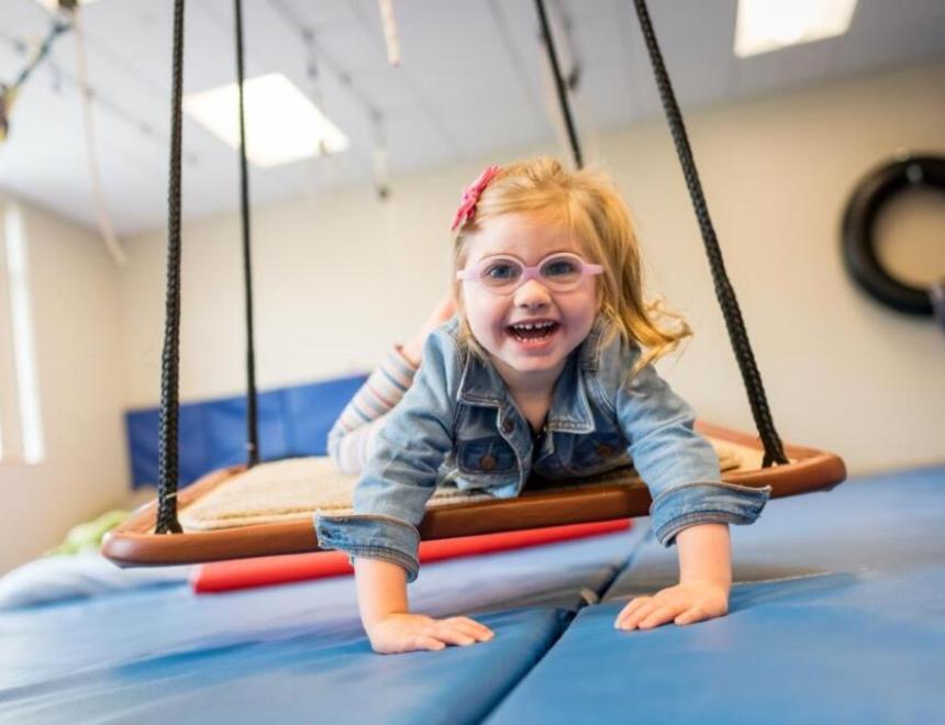 Little girl with blond pigtails and pink glasses swinging on a therapy swing in an OT gym.
