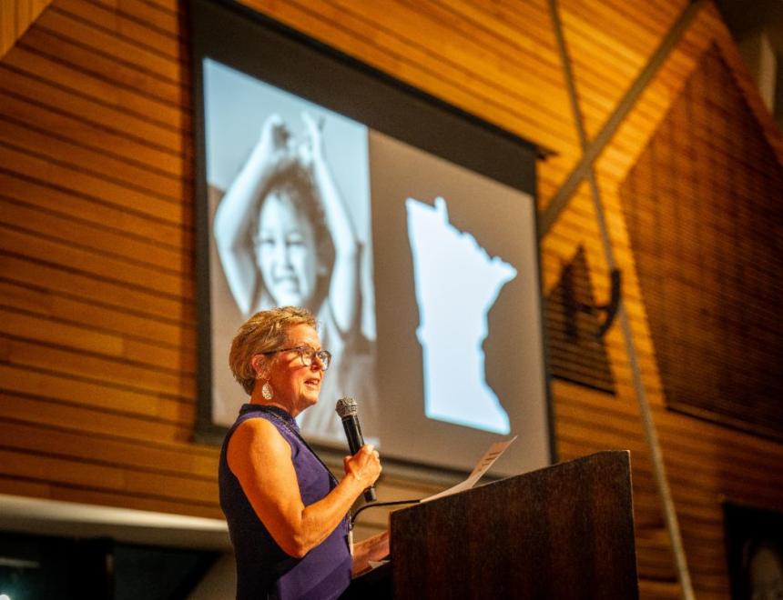 Woman with short blond hair speaks behind a podium on stage with a screen behind her depicting a photo of a happy child next to the outline of the state of Minnesota.