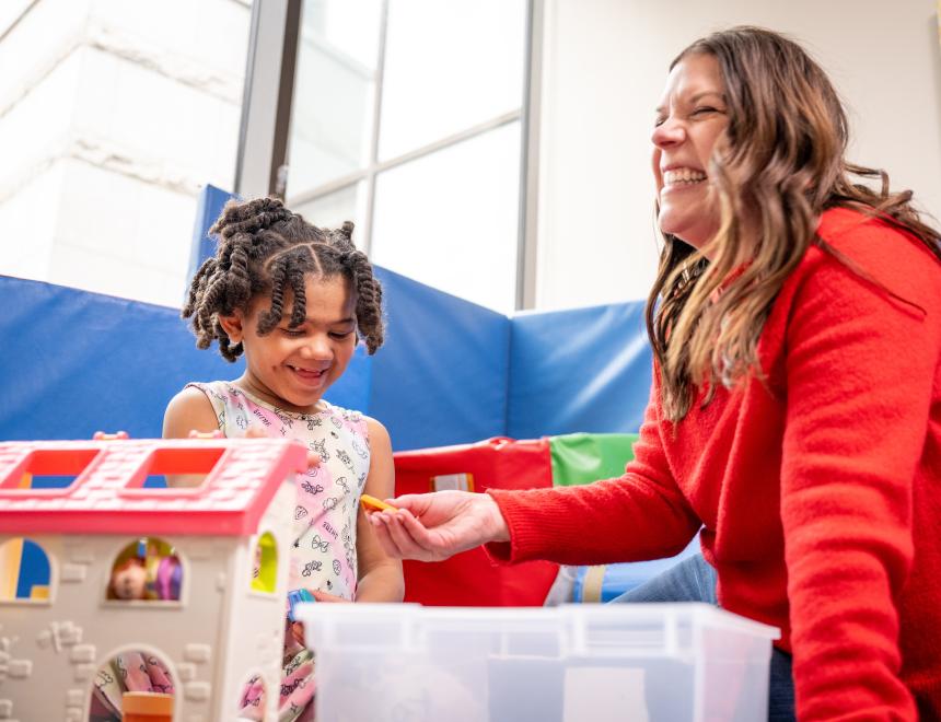 Little girl with dark skin and braids playing with toys alongside a woman wearing a red sweater.