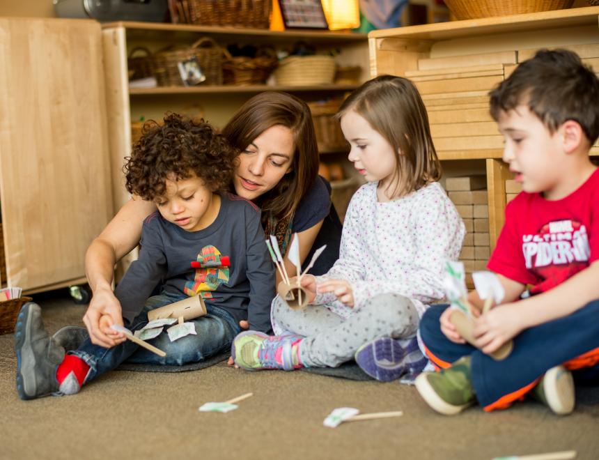 A teacher sits on the floor and interacts with three children, in a classroom.
