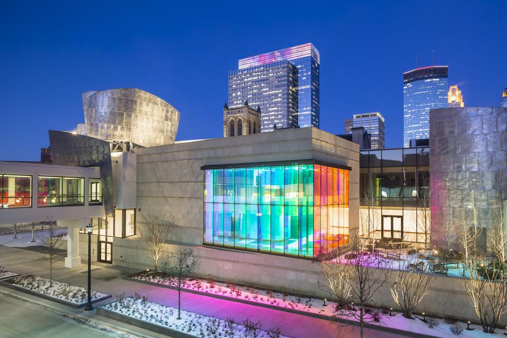 A nighttime photo of part of the downtown Minneapolis skyline featuring a building with internally lit windows casting a rainbow glow onto the sidewalk and street. 