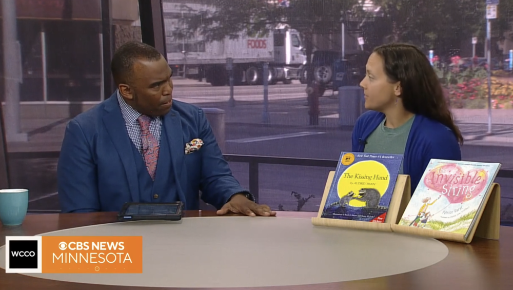 still image of a tv news desk with an interviewer, interviewee and children's book displayed