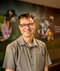 White man with short brown hair and glasses, wearing a grey button-up shirt, smiling.