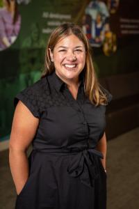 White woman with long brown hair, wearing a short-sleeved black dress with her hands in her pockets.