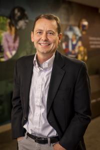 White man with brown hair, wearing a suite coat and button-up shirt, smiling with his hands in his pockets.