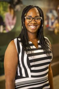 Dark-skinned woman with long braids, wearing glasses and a black and white striped dress, smiling. 