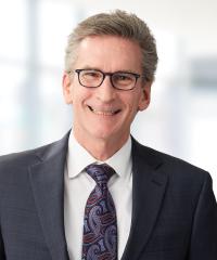 Smiling older white gentleman wearing glasses, a dark suit, light shirt and tie. 