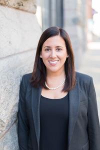 White woman with shoulder-length brown hair, wearing a grey suit jacket and black top, smiling.