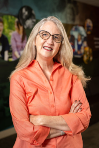 White woman with long blonde hair, wearing glasses and a coral shirt. 