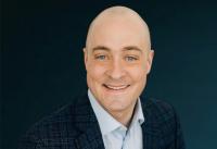 White man with no hair wearing a button-up shirt and suit jacket, smiling.