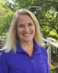 Blonde woman wearing a purple zip-up top, smiling, with green trees behind her. 