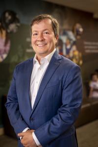 Man with tan skin and brown hair, wearing a blue suit jacket and light colored button-up shirt, smiling.