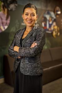 Woman with tan skin and pulled back dark hair, wearing a suit jacket and skirt, smiling.