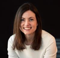 White woman with long brown hair, wearing a cream long-sleeved top, smiling.