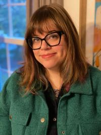 White woman with bangs and long brown hair, thick black glasses and green top, smiling.