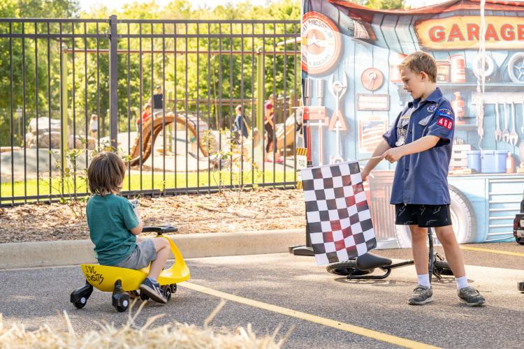 One child riding a toy vehicle while another child holds out a black and white race flag.