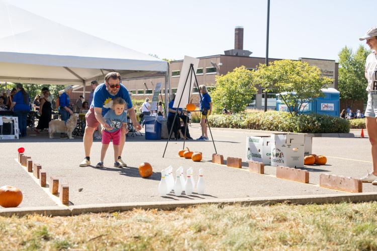Dad and daughter try to knock bowling pins down by rolling a pumpkin.