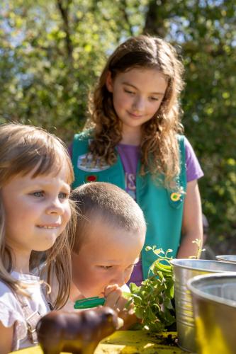 A girl scout helps two younger kids plant herbs in pots.