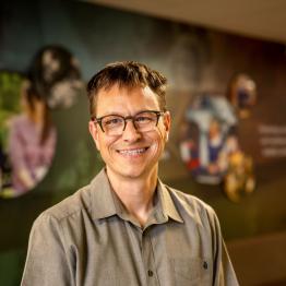White man with short brown hair and glasses, wearing a grey button-up shirt, smiling.