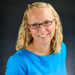 White female blonde woman wearing a bright blue top, smiling.