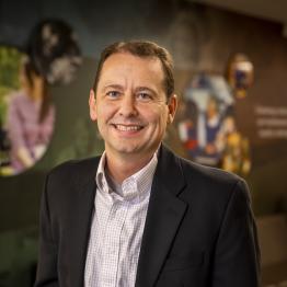 White man with brown hair, wearing a suite coat and button-up shirt, smiling with his hands in his pockets.