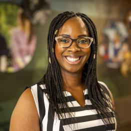 Dark-skinned woman with long braids, wearing glasses and a black and white striped dress, smiling. 