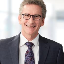 Smiling older white gentleman wearing glasses, a dark suit, light shirt and tie. 