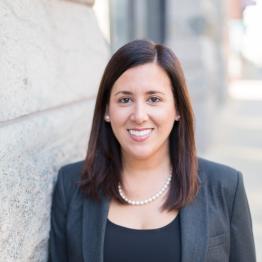 White woman with shoulder-length brown hair, wearing a grey suit jacket and black top, smiling.