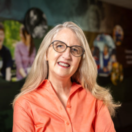 White woman with long blonde hair, wearing glasses and a coral shirt. 