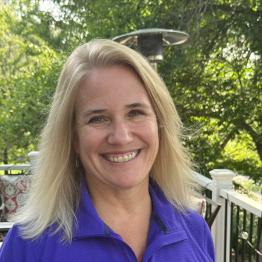 Blonde woman wearing a purple zip-up top, smiling, with green trees behind her. 