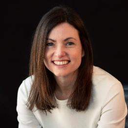 White woman with long brown hair, wearing a cream long-sleeved top, smiling.