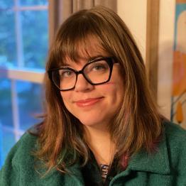 White woman with bangs and long brown hair, thick black glasses and green top, smiling.