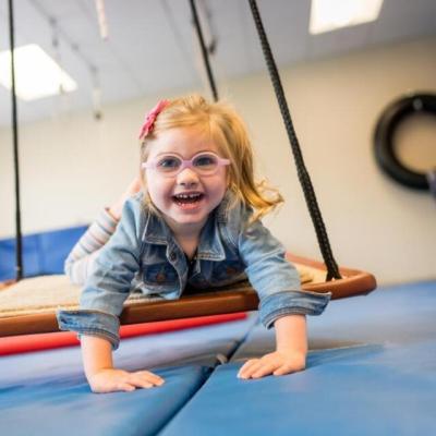 Little girl with blond pigtails and pink glasses swinging on a therapy swing in an OT gym.