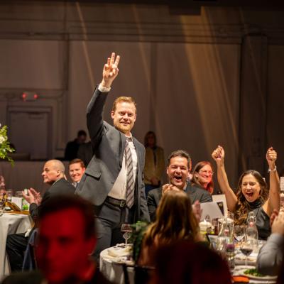 a group of adults at a dinner gala, with one person standing, raising his hand.