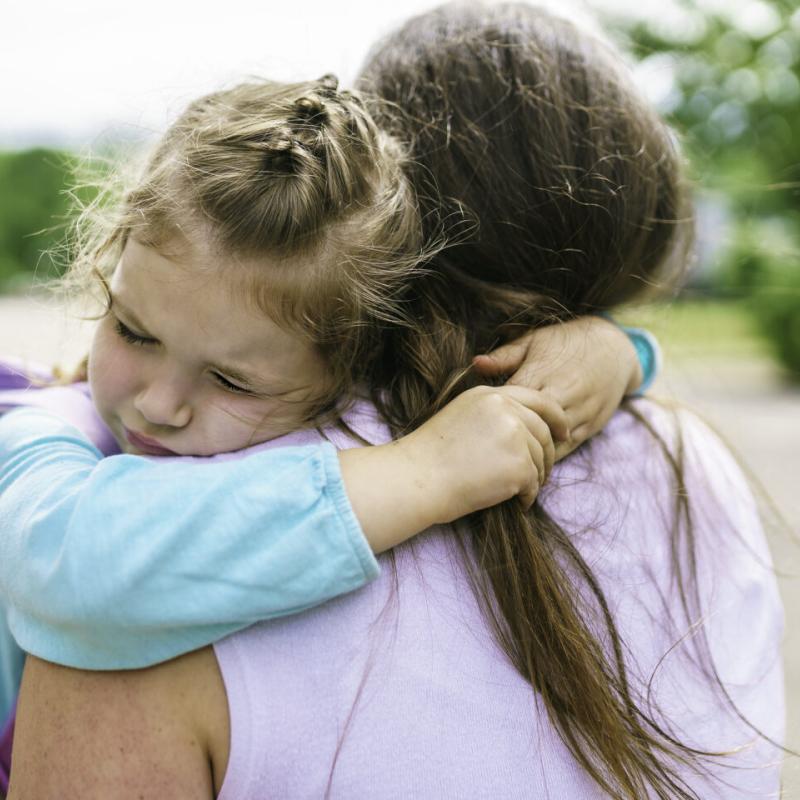child hugging adult woman; woman is shown from behind. Child is wearing a backpack.