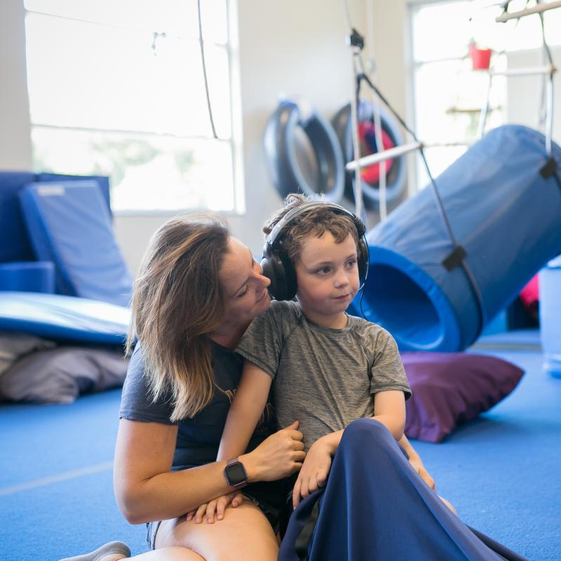 A female caregiver and child, wearing noise-blocking headphones, sit together in an occupational therapy gym on blue foam mats. 